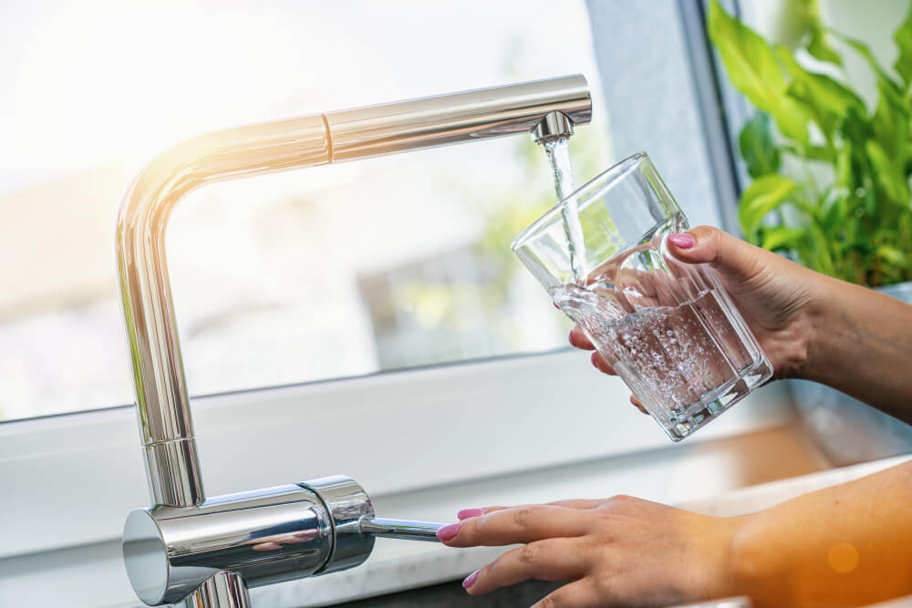 Person filling a clear glass with water from a modern chrome kitchen faucet near a window with green plants.