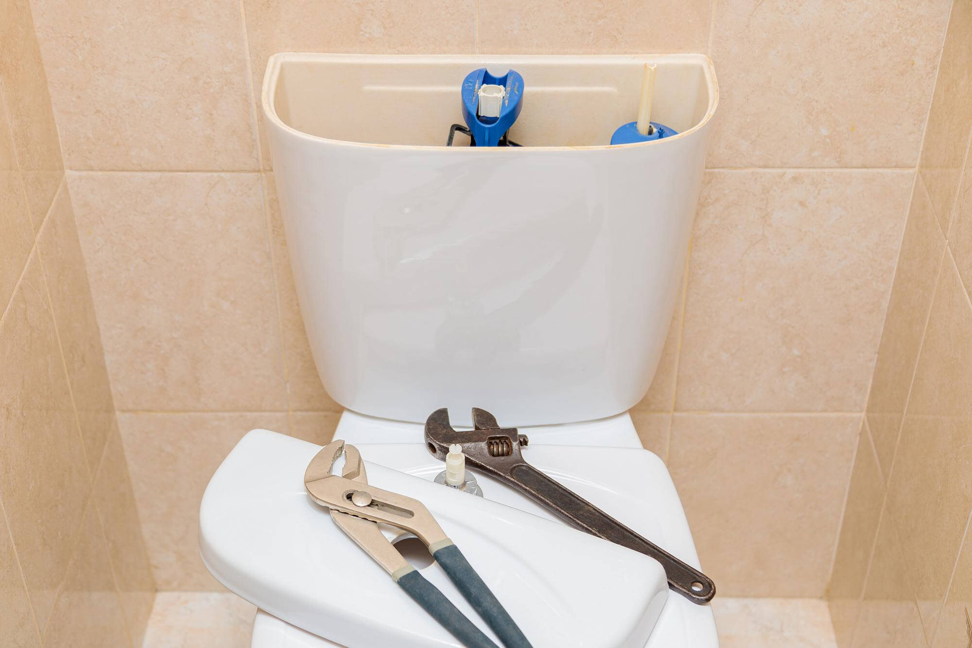 Open toilet tank with tools including adjustable wrench and slip-joint pliers on the closed toilet lid.