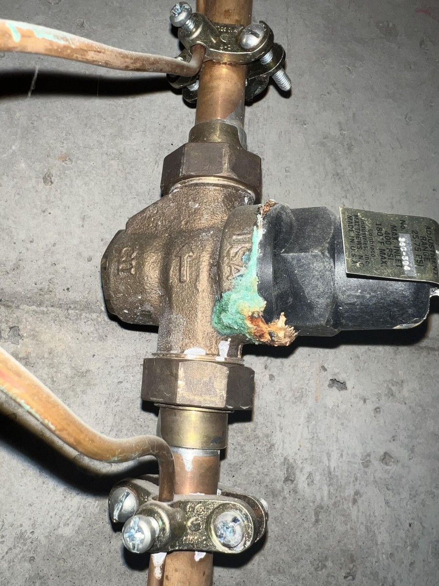 Close-up of a corroded brass valve connected to copper pipes with clamps on a concrete wall.