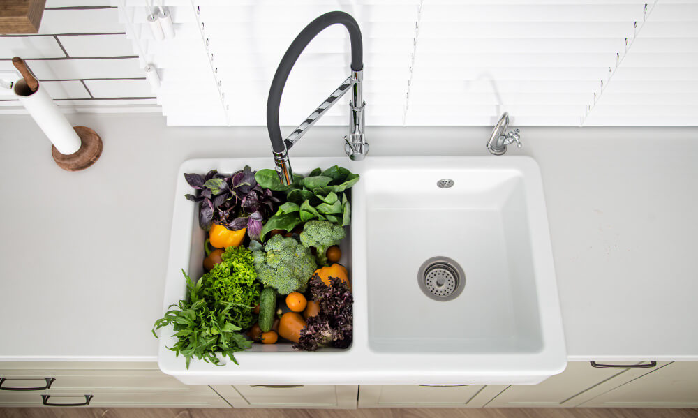 Top view of a white double kitchen sink filled with fresh vegetables including broccoli, lettuce, cucumbers, and bell peppers, with a modern black and chrome faucet.