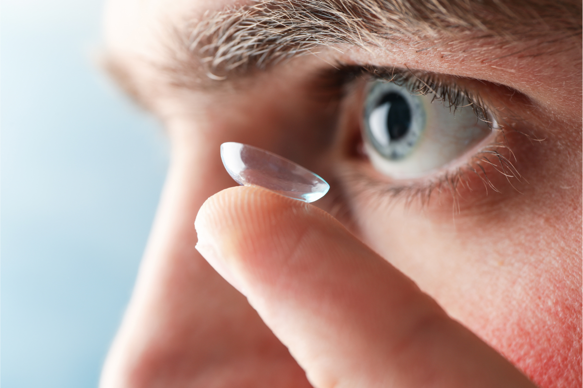 Close-up of a man with blue eyes holding a contact lens on his fingertip near his eye.