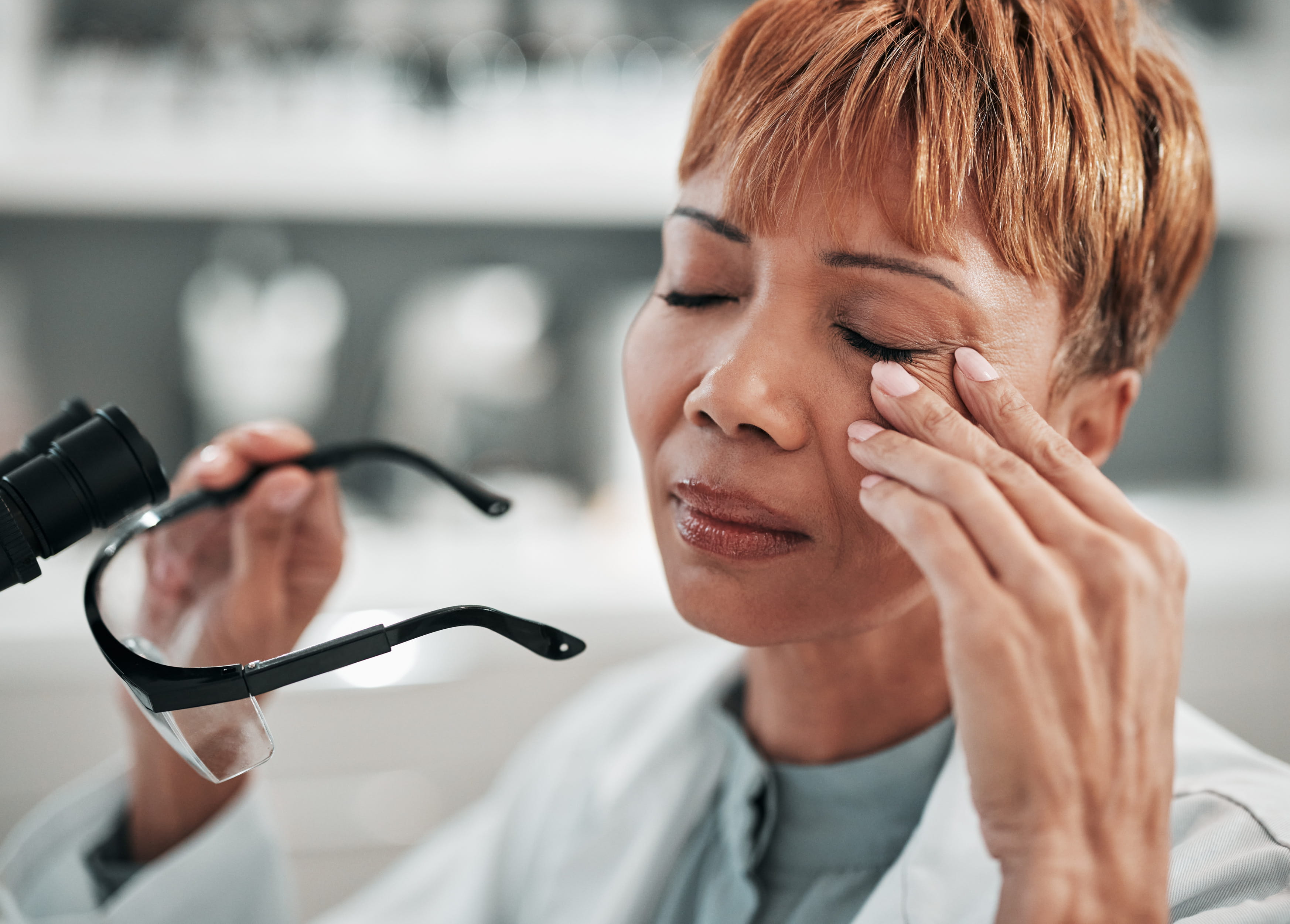 Woman with reddish hair removing safety glasses, touching face with discomfort expression.
