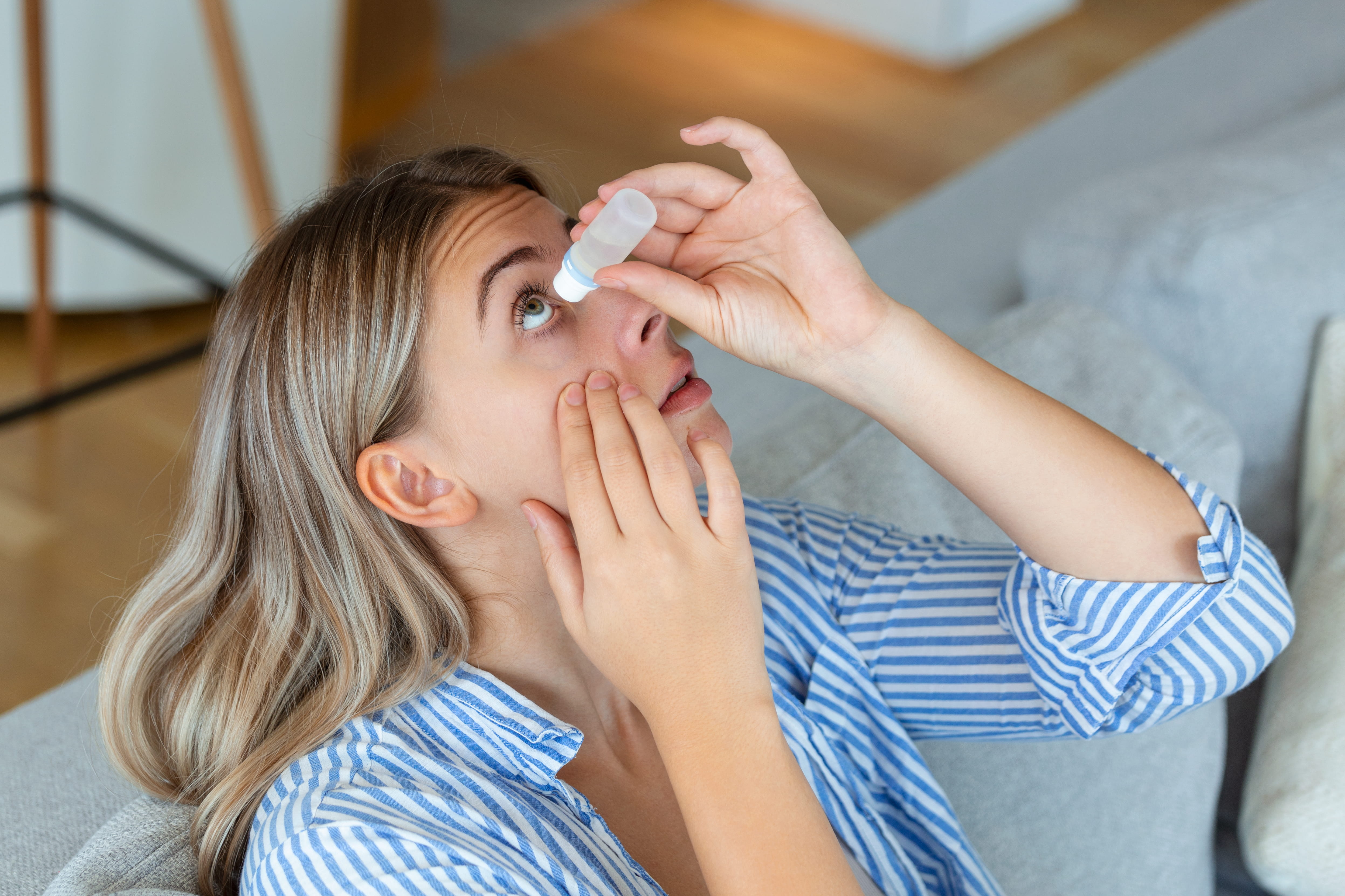 Woman applying eye drops to her eye while sitting on couch indoors
