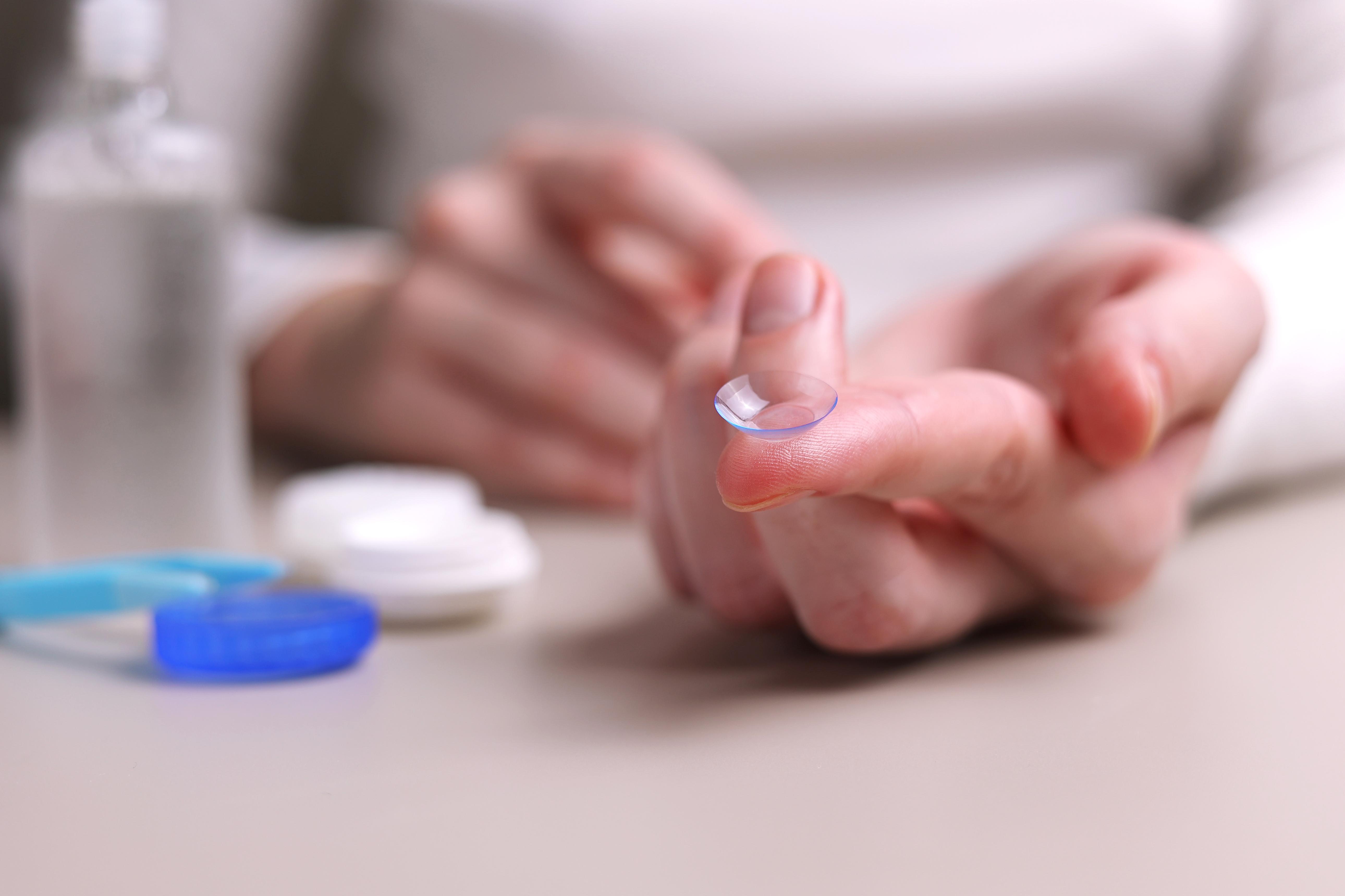 Person holding a contact lens on their fingertip with care supplies in background