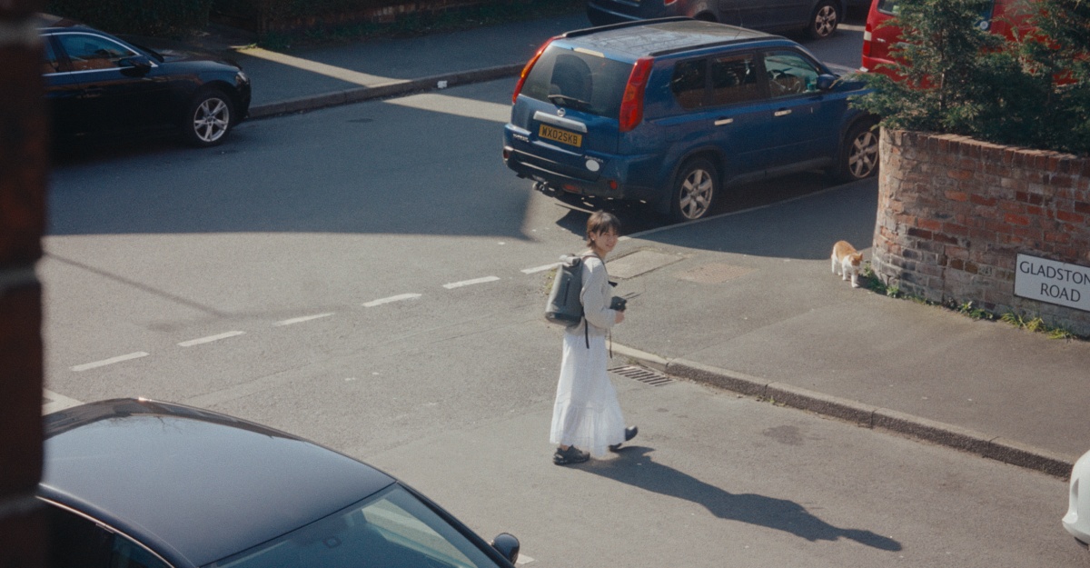 Person in white skirt crossing a street near parked cars and a brick wall with a cat nearby.