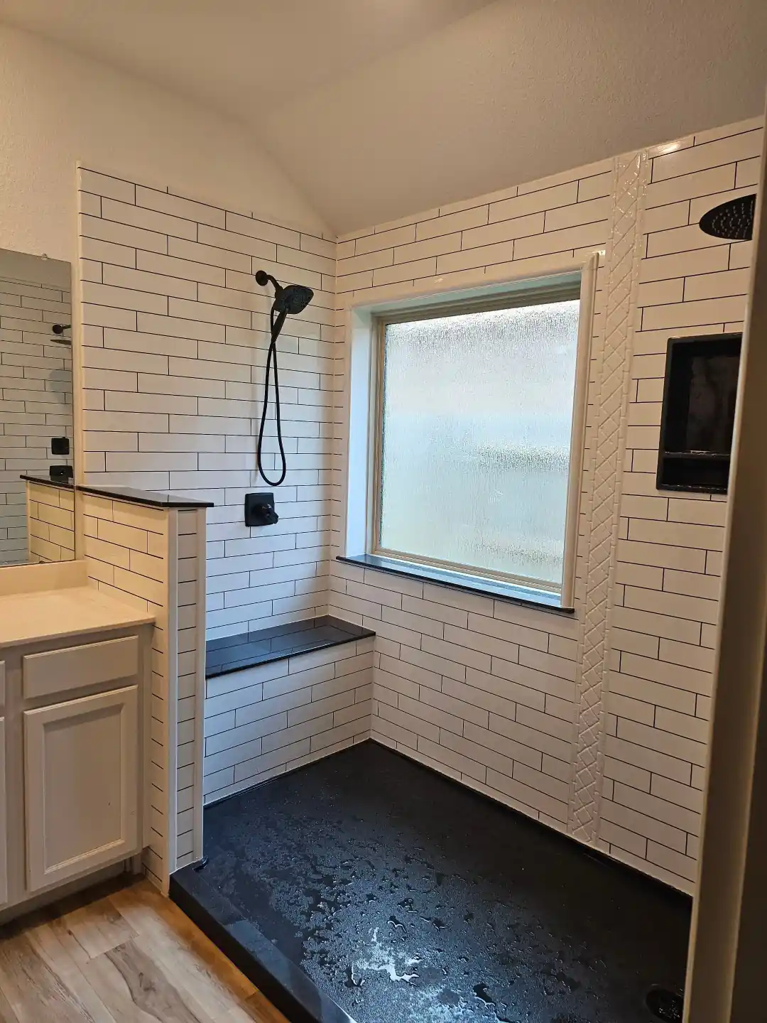 A contemporary walk-in shower with white subway tile walls, a built-in black bench, matte black handheld and rain showerheads, and a frosted window providing natural light, combining modern style with practical comfort.