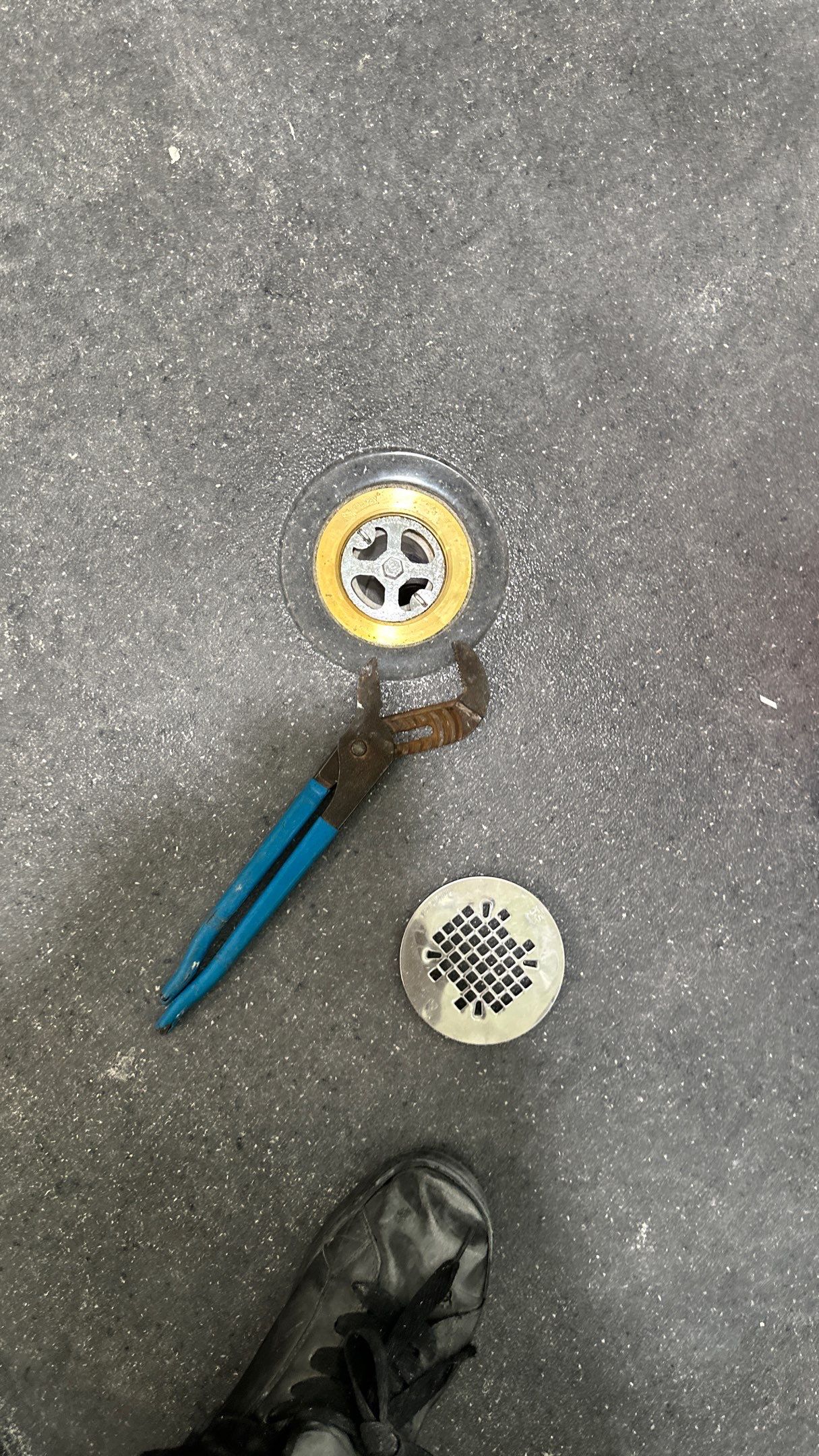 A close-up of a shower drain with the cover removed, a pair of blue-handled adjustable pliers gripping the drain ring, and a worker’s black shoe visible on the bathroom floor.