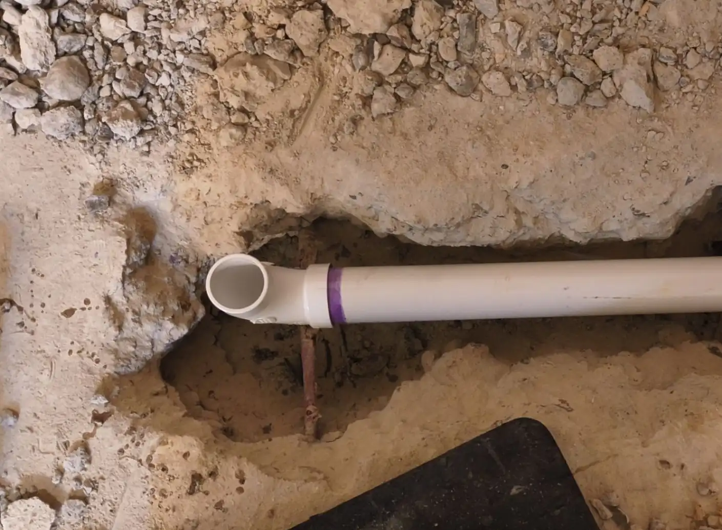 A white PVC pipe with a newly attached elbow joint lies in a trench cut through a concrete slab. Directly beneath the pipe is a post-tension cable embedded in the foundation, showing the potential risks if accidentally damaged during plumbing work.