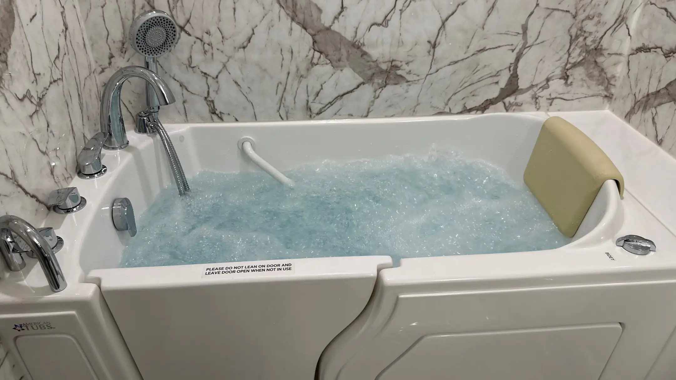 A white walk-in tub with bubbling blue water, chrome faucet controls, a handheld showerhead, and a beige headrest, set against a wall with a marble-look finish, providing a spa-like experience with easy access.