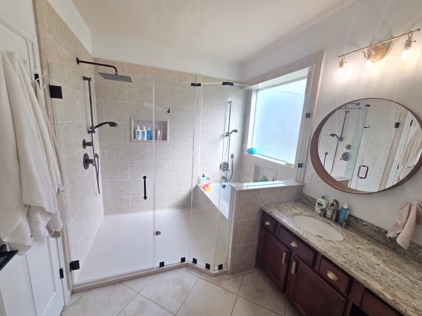 A modern bathroom with a large glass-enclosed walk-in shower featuring dual black showerheads and beige tile walls. The shower includes built-in shelves with bottles and accessories, and a large frosted window allows natural light in. To the right, there's a granite countertop vanity with a round mirror and brushed bronze light fixture above.
