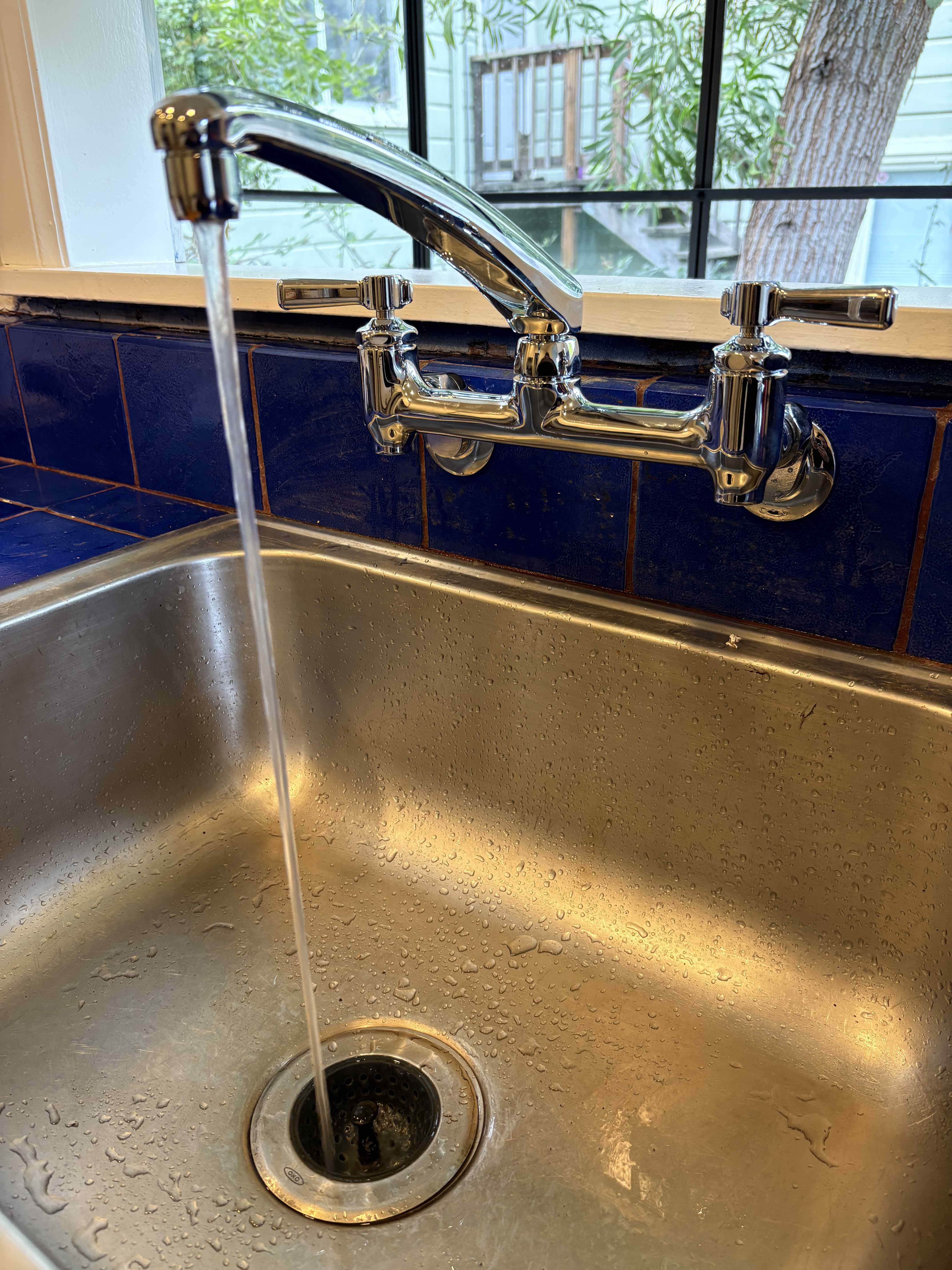 A modern bathroom featuring a glass-enclosed walk-in shower with rain showerhead on the left, a streamlined white toilet centered under a window, and a floating wooden-topped vanity with two vessel sinks and chrome faucets on the right.