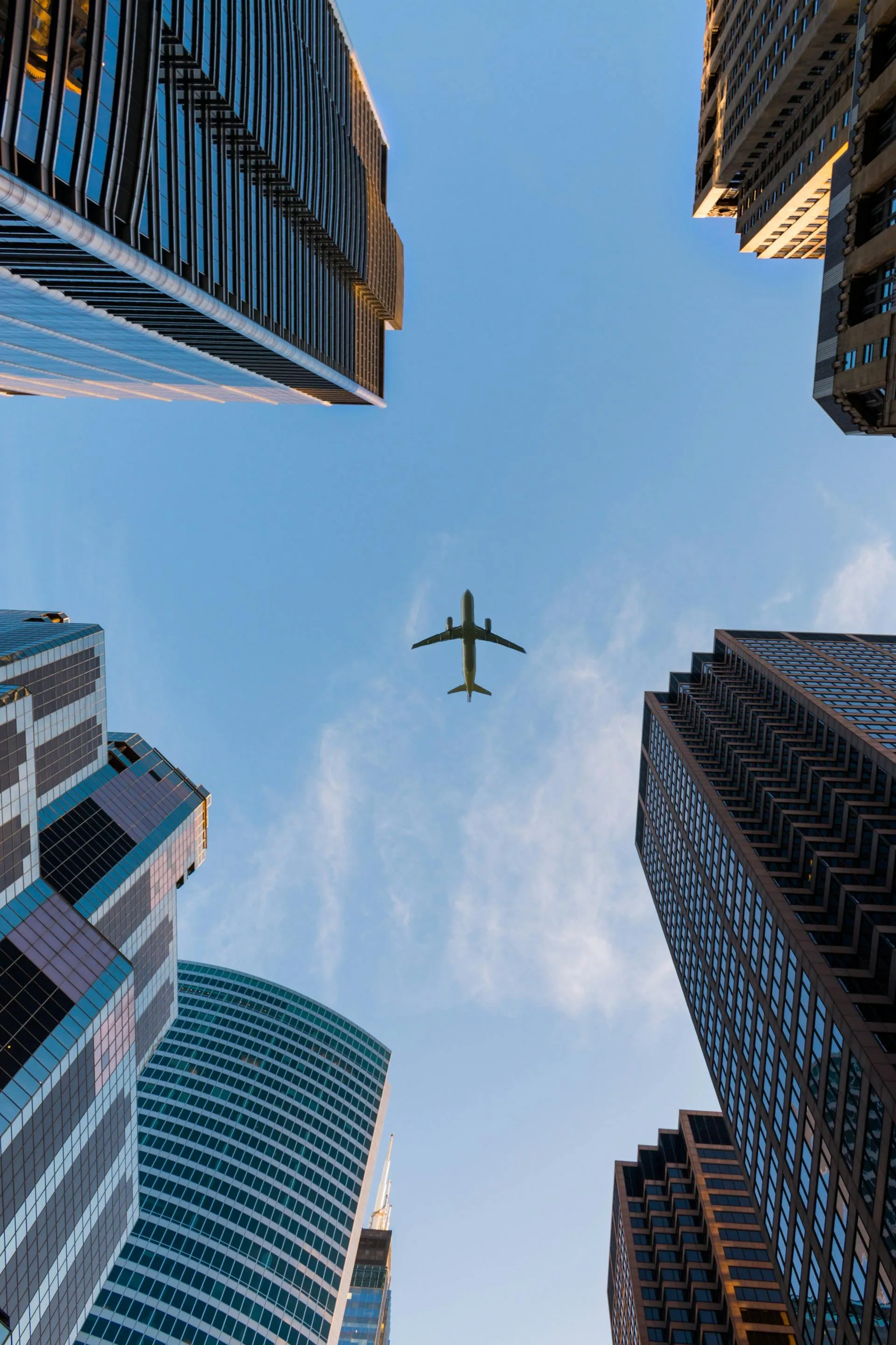 Blick nach oben zwischen mehreren Hochhäusern; ein Flugzeug fliegt zentral am blauen Himmel vorbei.