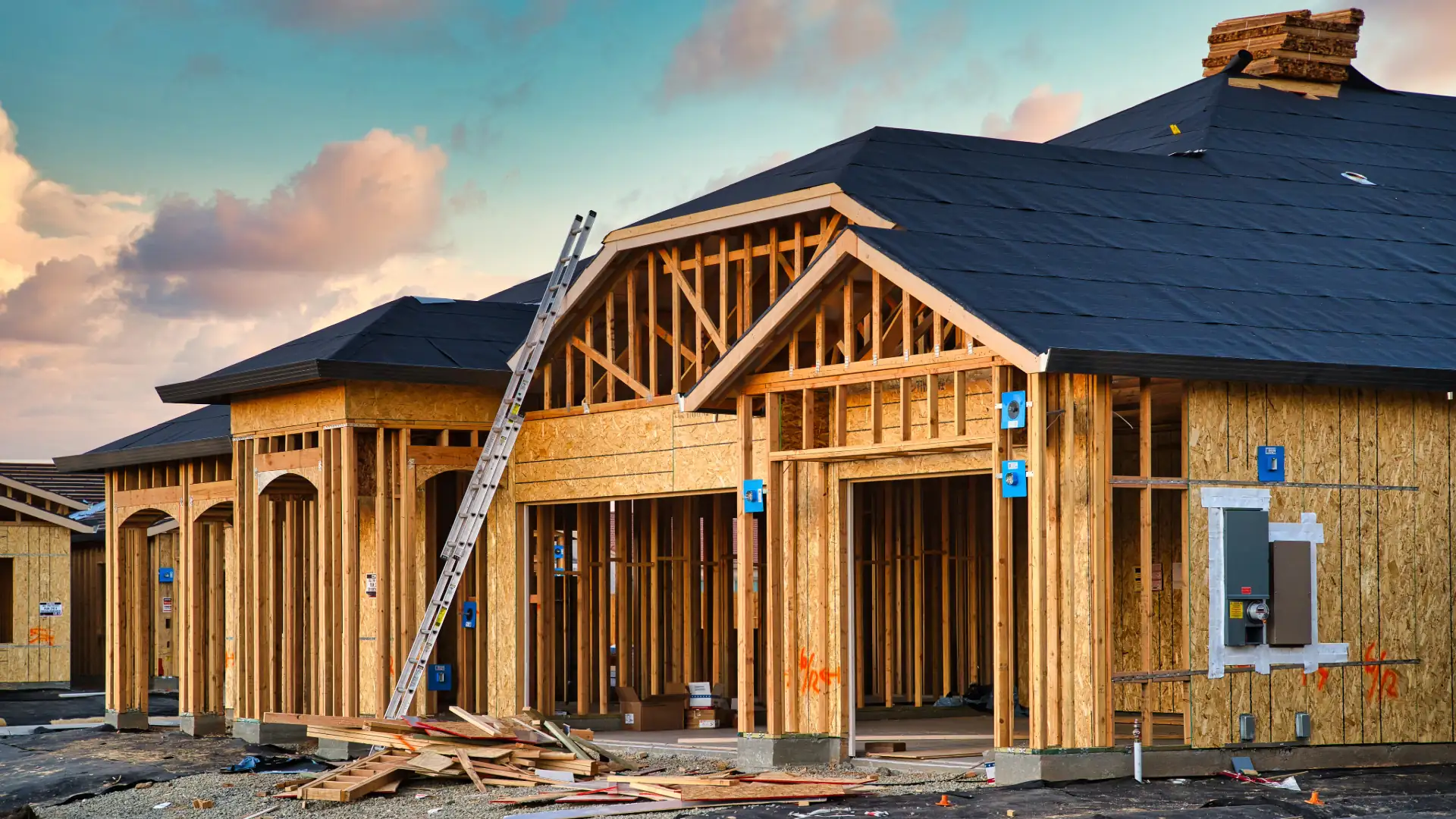 Framed and sheathed homes under construction with a ladder and lumber on site under a pink and teal evening sky