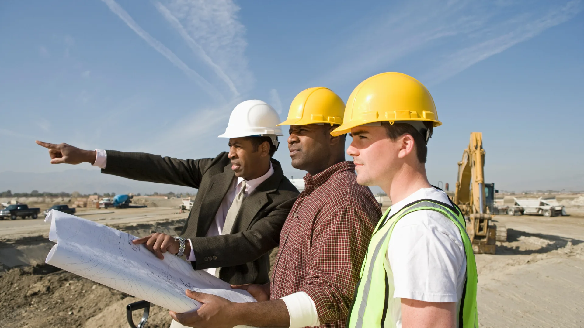 Surveyor and builders in hard hats reviewing plans and pointing across a construction site