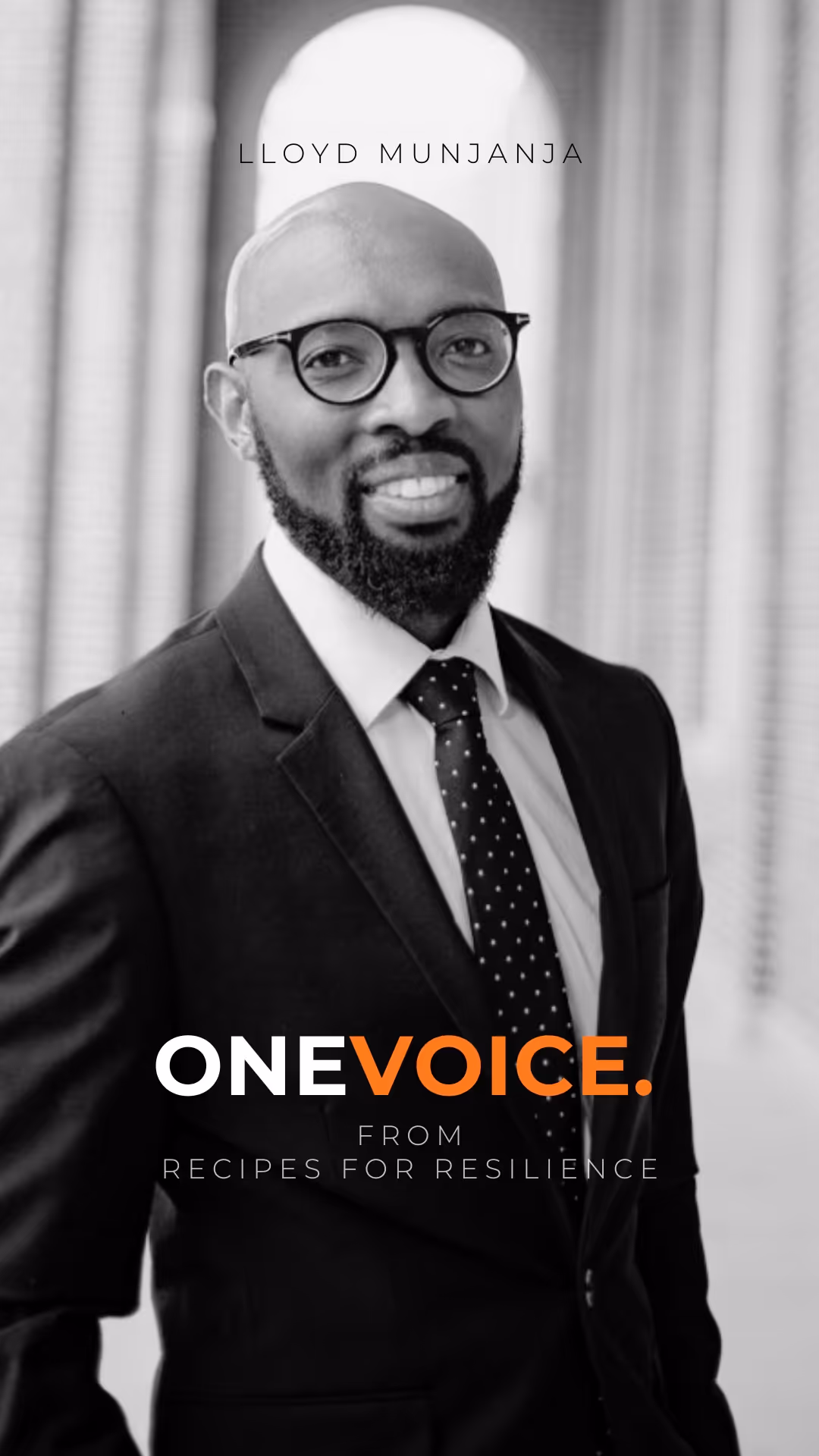 Black and white portrait of a smiling man with glasses and a beard wearing a suit and polka-dot tie in an elegant setting.