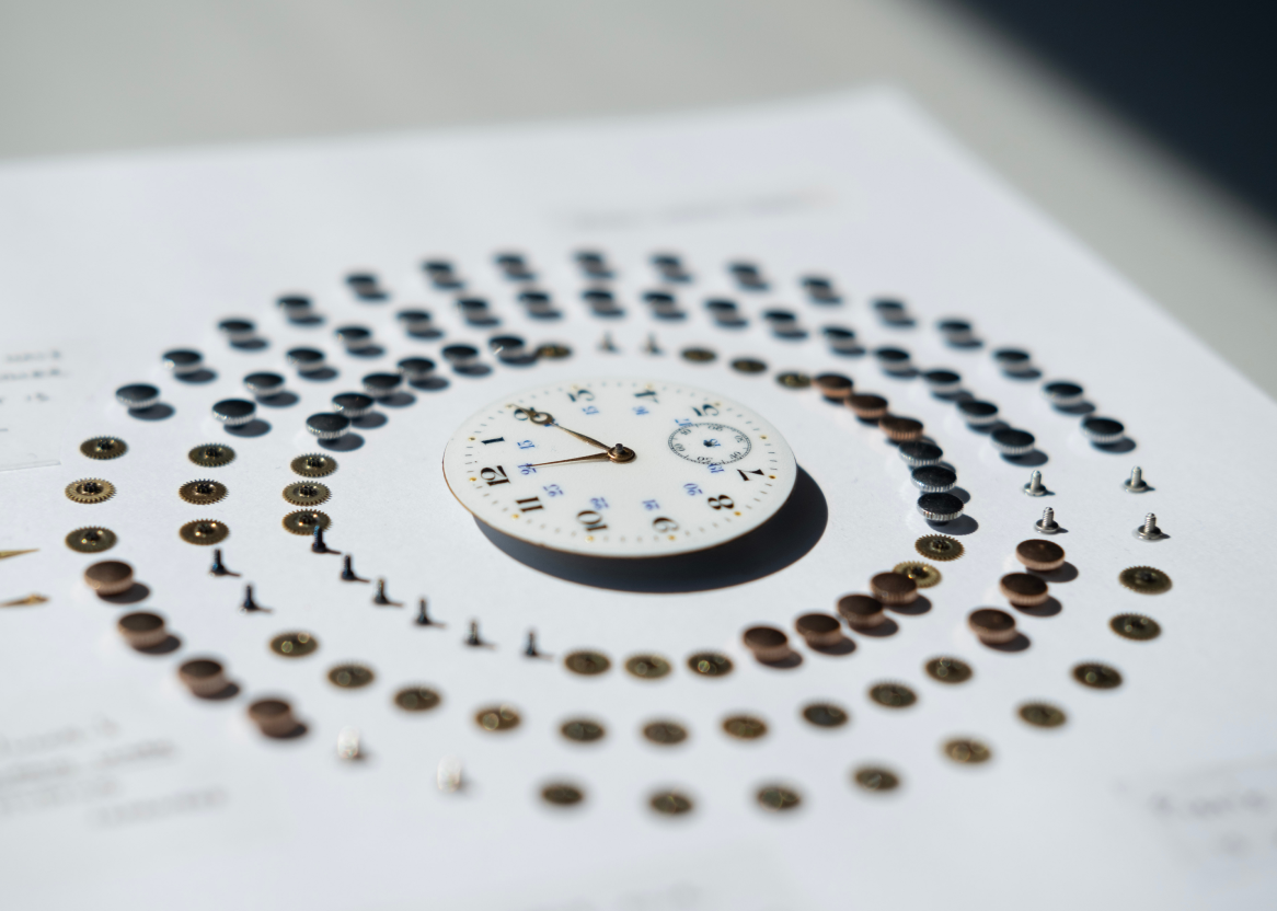 A closup of a white watch face with a modern look with bold black and tiny blue numbering. Around it run three circles of five types of watch parts corresponding to different activities, turning it into a data visualization about the three authors daily schedules and time.
