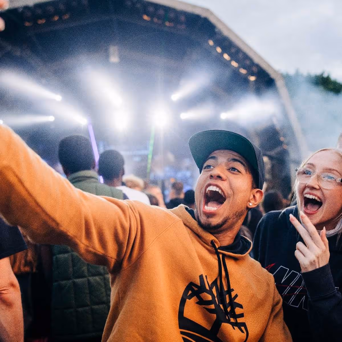 An energetic photo of two people smiling and taking a selfie at a festival  with stage lights in the background