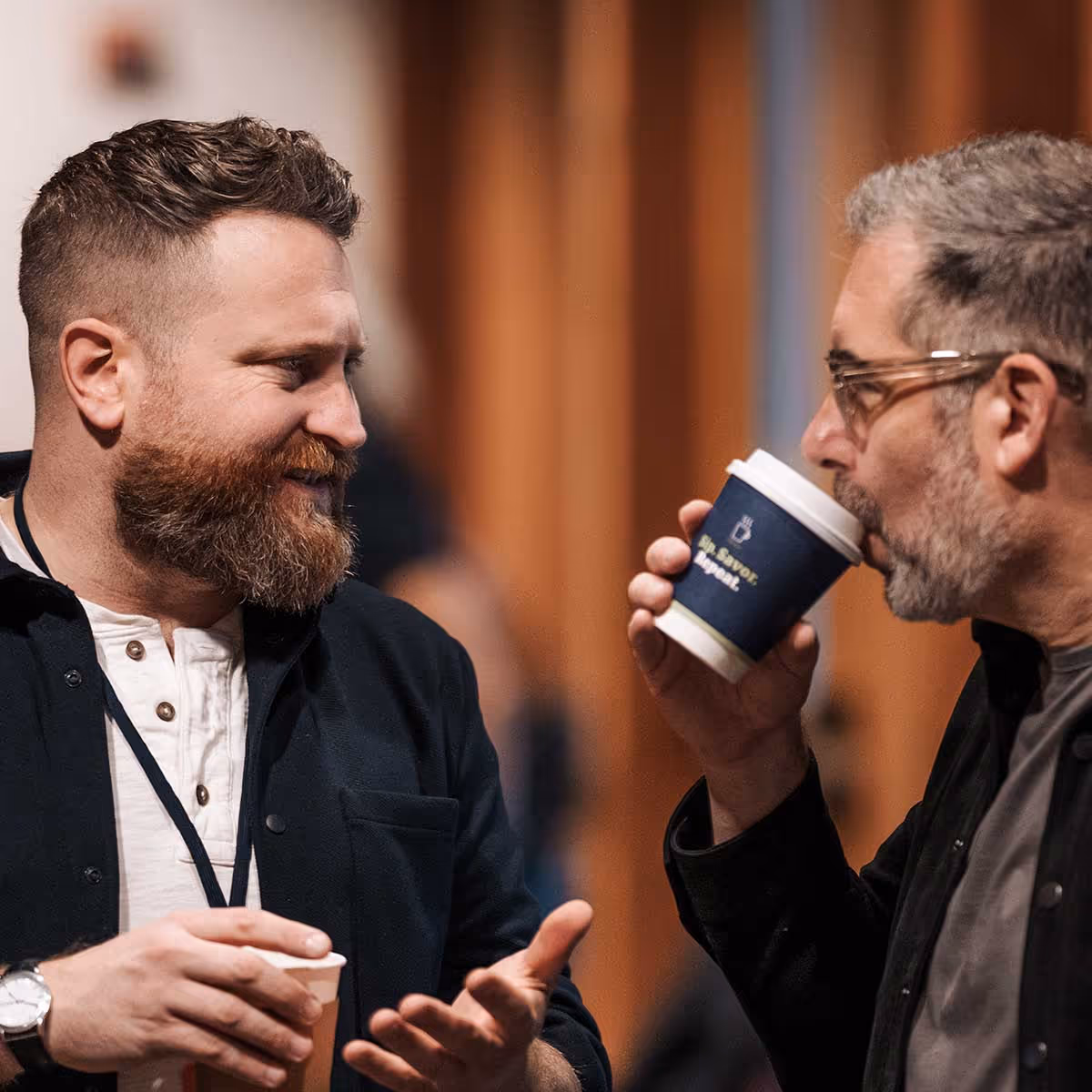 Two men smiling and talking to each other with coffee cups in their hands 