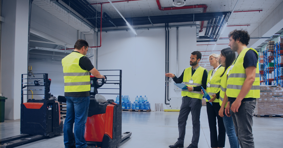 Warehouse workers receiving forklift safety training on the warehouse floor, with a supervisor demonstrating correct operating procedures.
