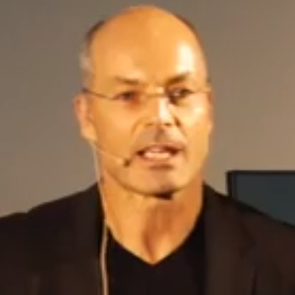 Man with glasses and bald head wearing a black jacket speaking with a headset microphone.