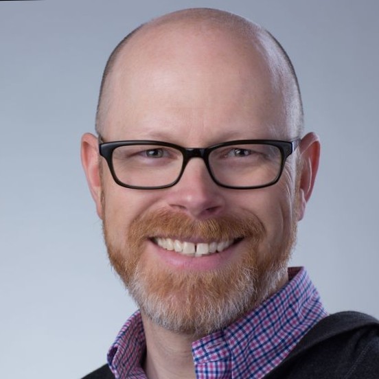 Smiling bald man with glasses, a beard, and a checkered shirt against a plain gray background.