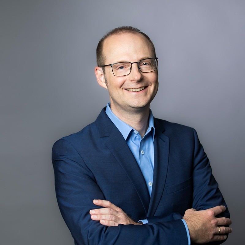 Smiling man in glasses wearing a blue suit and light blue shirt with arms crossed against gray background.