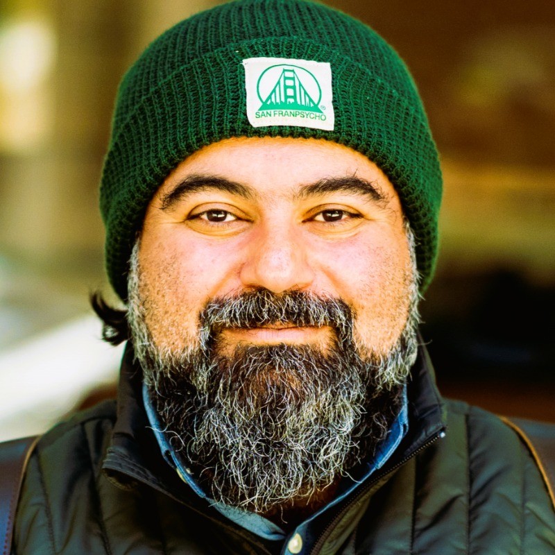 Bearded man wearing a green beanie with a San Franpsycho logo and a dark jacket, smiling at the camera.