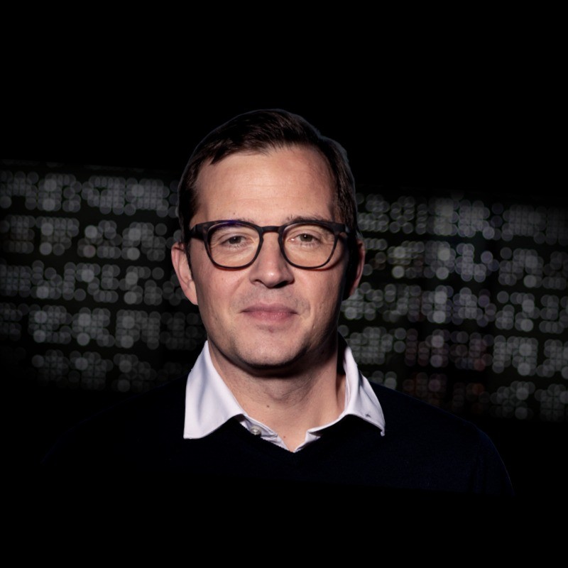 Man with glasses wearing a white collared shirt and dark sweater against a dark blurred background.