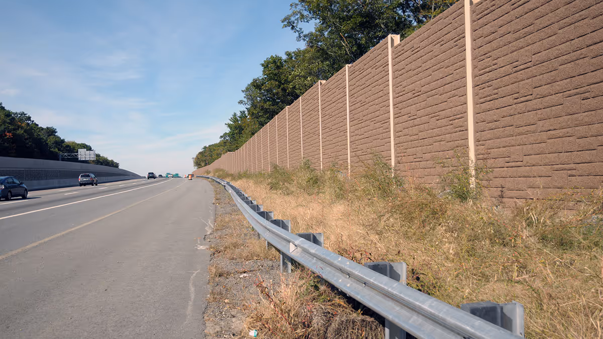 A highway with a metal guardrail runs beside a tall brown sound barrier wall and sparse vegetation.