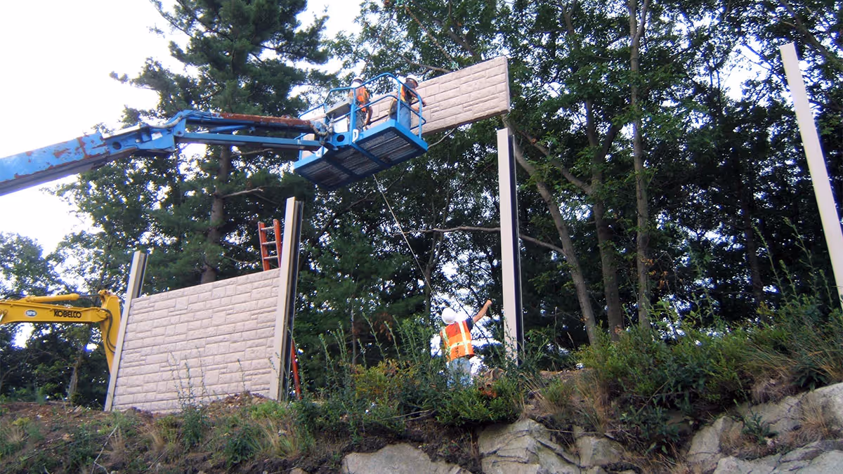 Workers on a lift install large wooden panels on tall posts outdoors near trees and rocks.