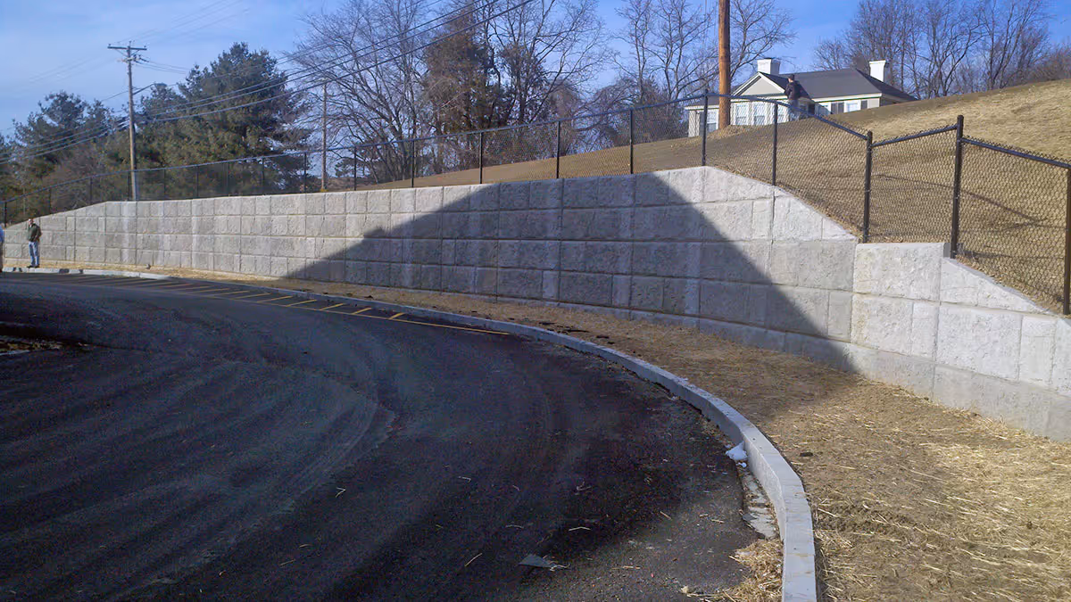 Concrete retaining wall along a curved road with a metal fence on top and a grassy slope beside it under a clear sky.