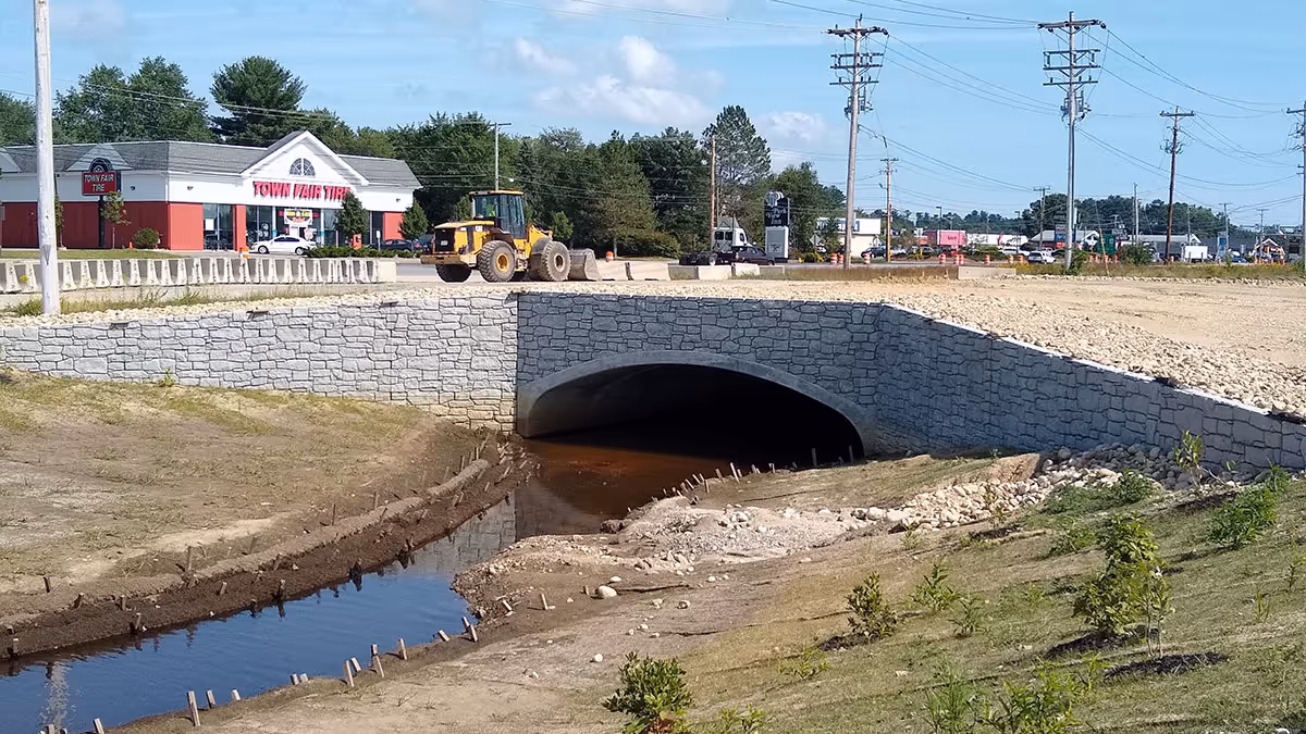 A stone bridge crosses over a narrow creek, with construction equipment and buildings visible in the background.