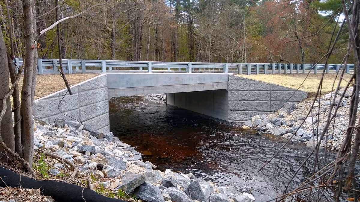 A small concrete bridge with metal railings spans over a rocky stream surrounded by trees and vegetation.