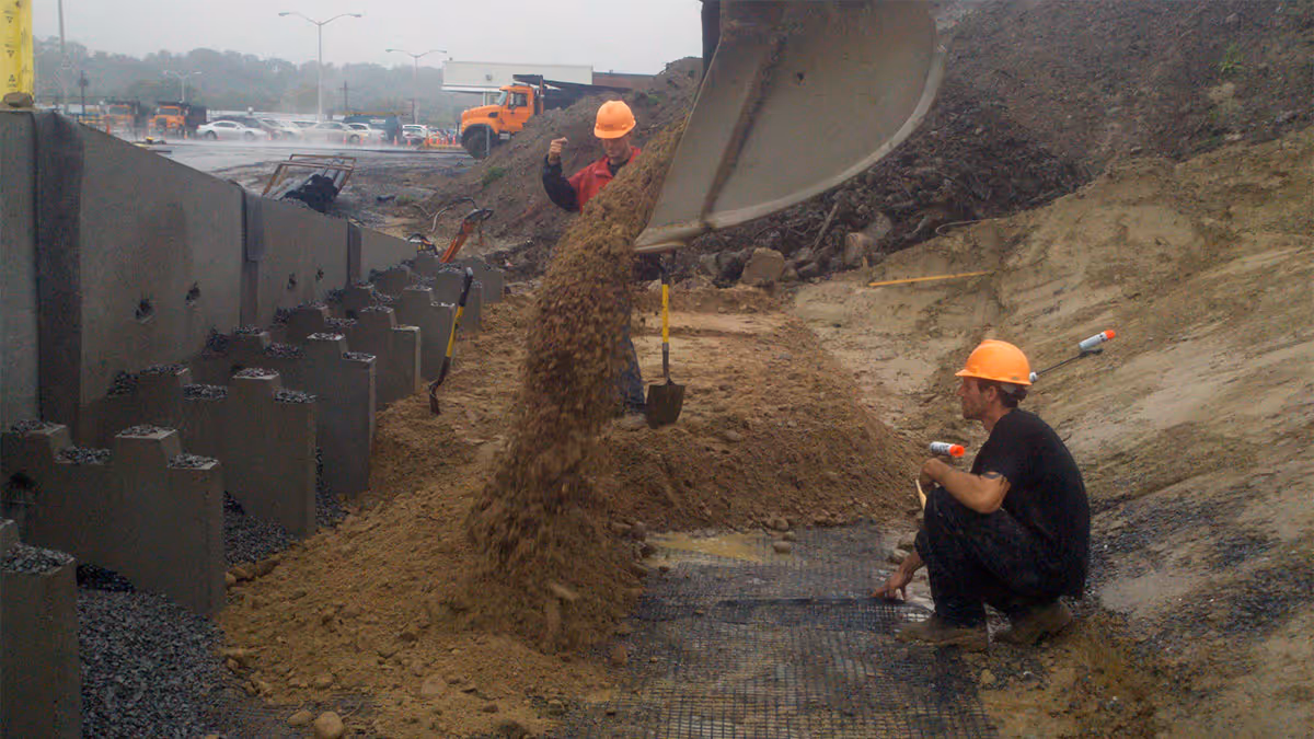 Two workers in orange helmets guide soil being dumped by a large machine onto a sloped construction site.