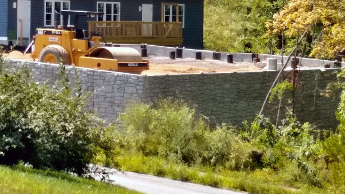 A bulldozer sits on a construction site near a green building, with grass and shrubs in the foreground.