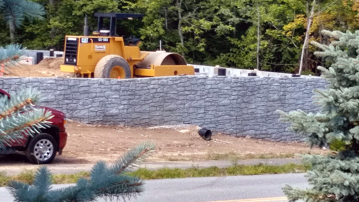 A yellow road roller is parked on dirt behind a gray stone wall, with trees and a red car in the foreground.