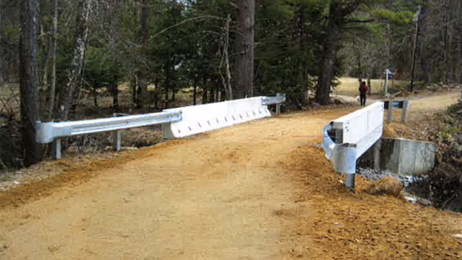 A narrow dirt road crosses a small bridge with white metal railings in a wooded area.