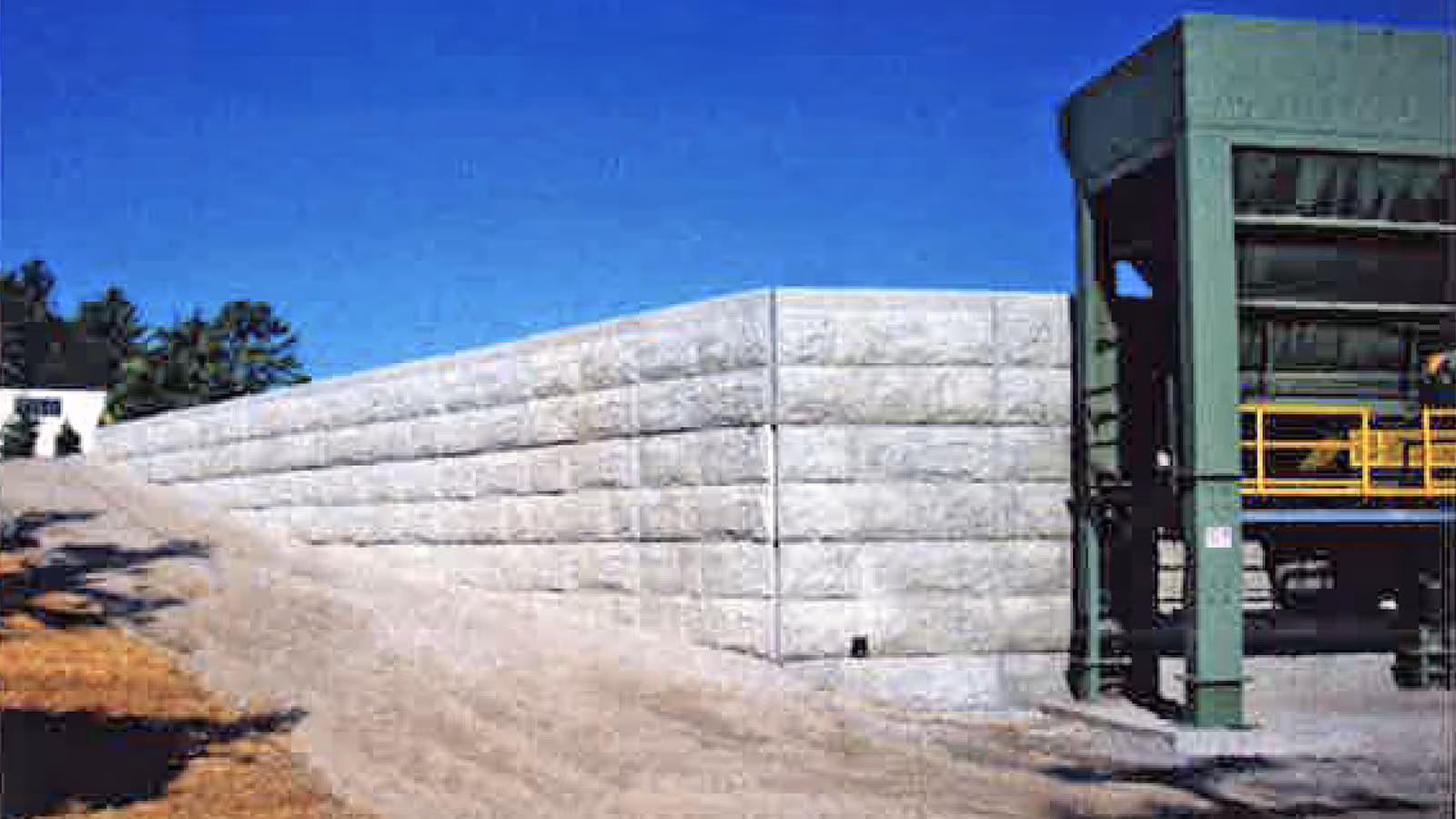 Large concrete retaining wall with a sloped driveway on the left and a green industrial structure on the right under a clear sky.