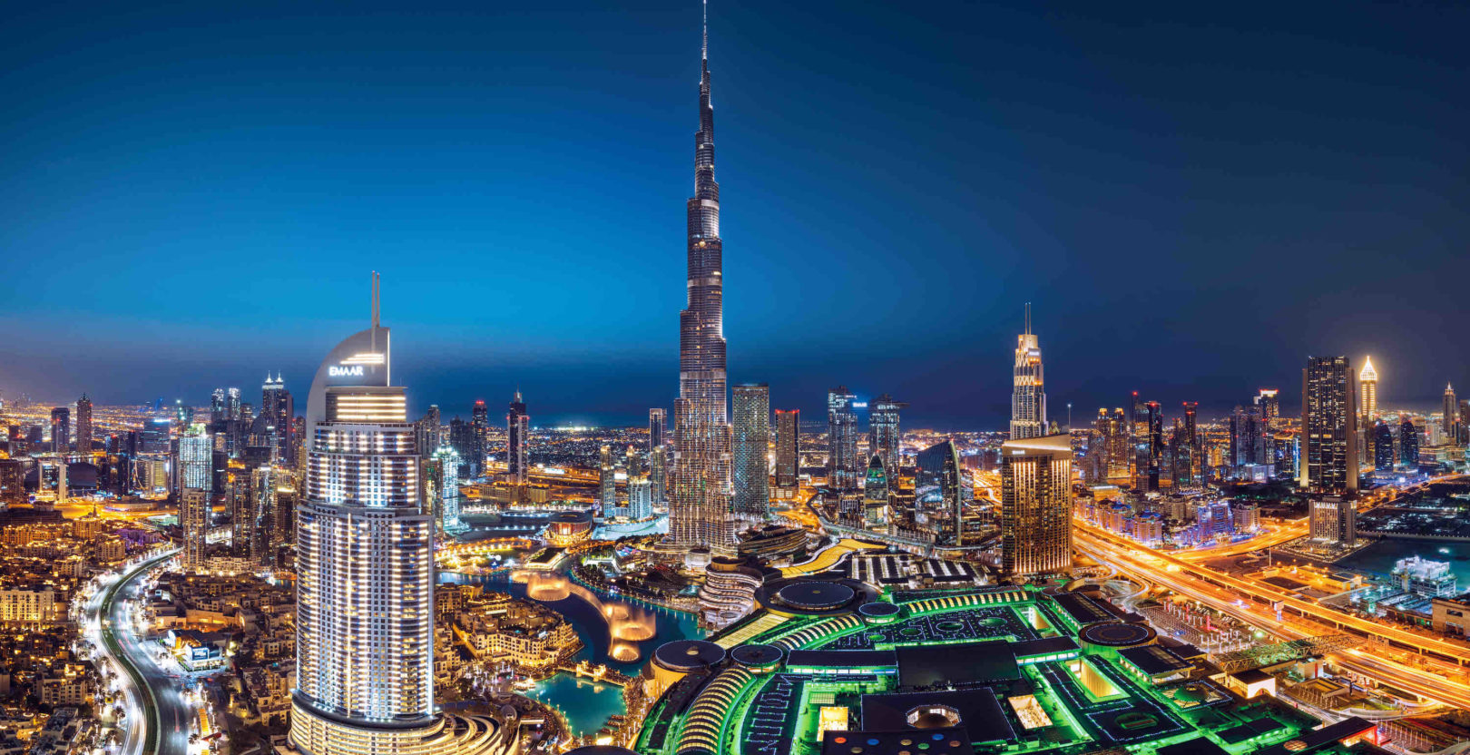Aerial view of the Dubai skyline at night featuring the illuminated Burj Khalifa, Emaar building, highways, and vibrant cityscape