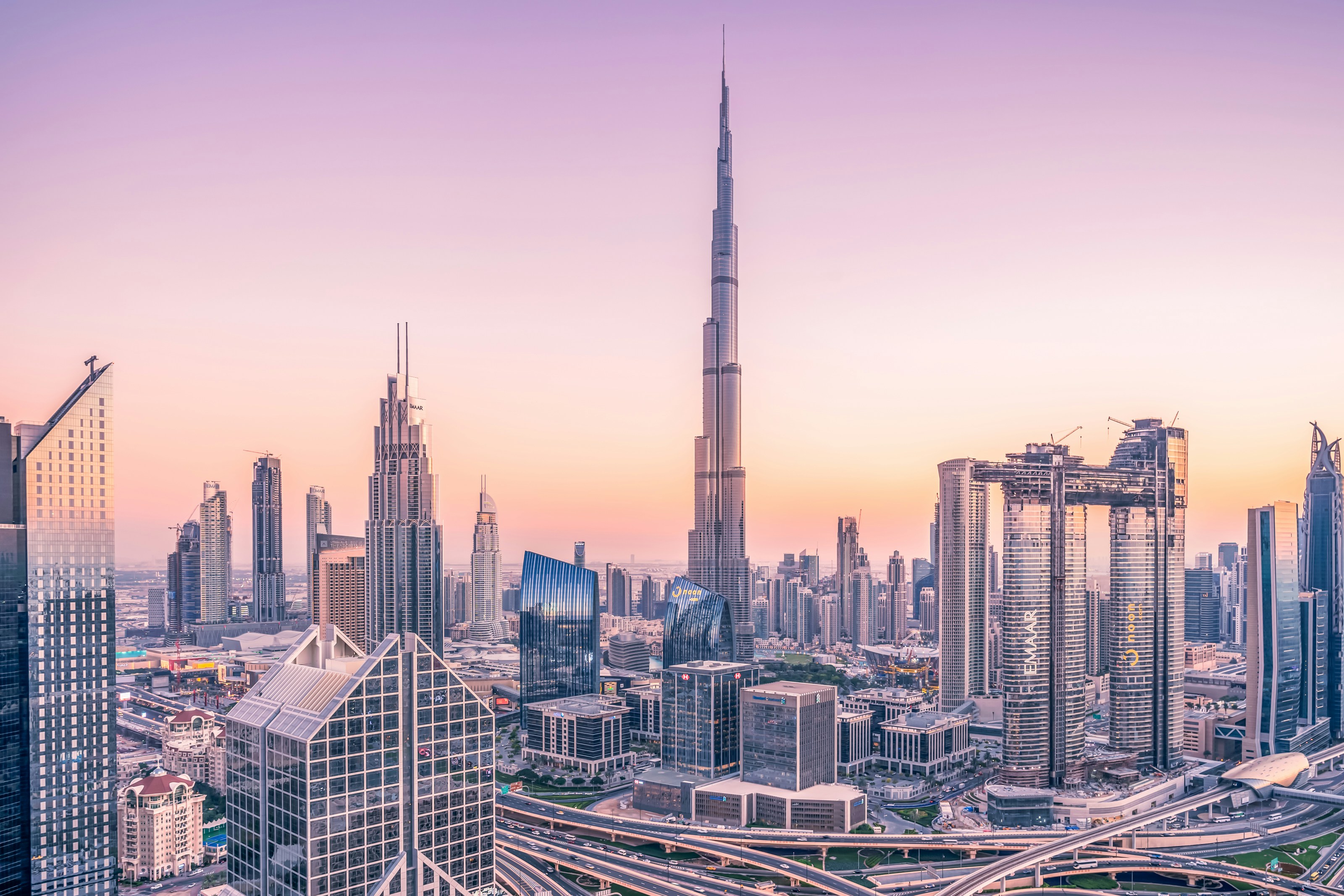 Dubai skyline at sunset featuring the Burj Khalifa, modern skyscrapers, and elevated highways under a pastel sky