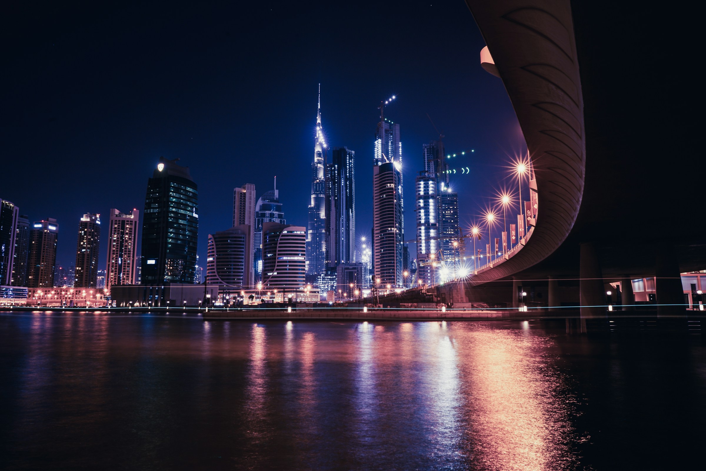 Dubai skyline at night with illuminated skyscrapers, including the Burj Khalifa, reflecting on the water and a modern bridge in the foreground