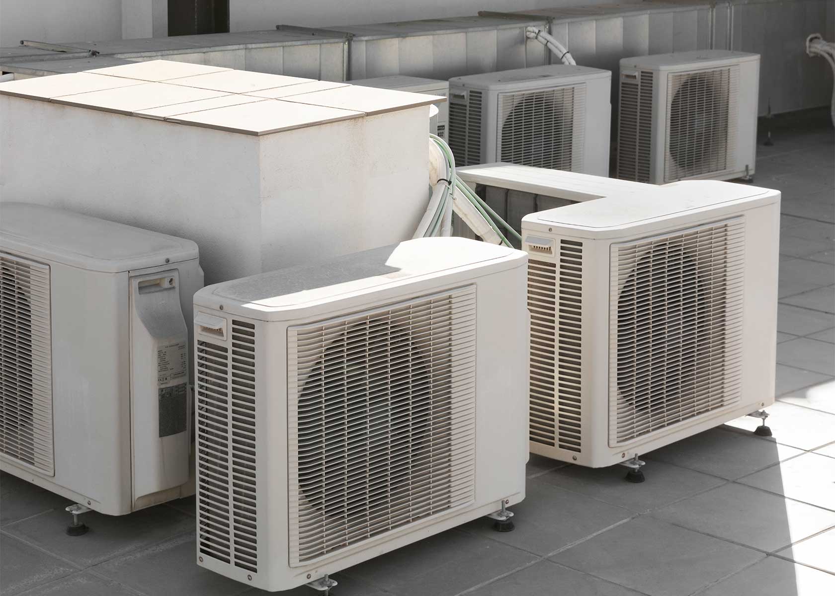 Outdoor units of heat pumps and air conditioning systems installed on a building rooftop with residential apartments in the background.