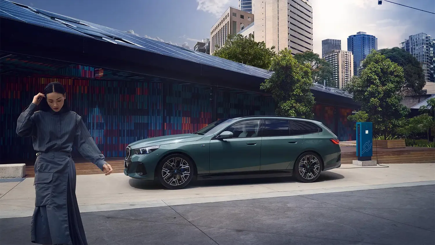 BMW i5 Touring in grün-metallic beim Laden an einer öffentlichen Ladesäule vor urbaner Kulisse – BMW Wolter & Steiner in Neubrandenburg, Waren und Prenzlau