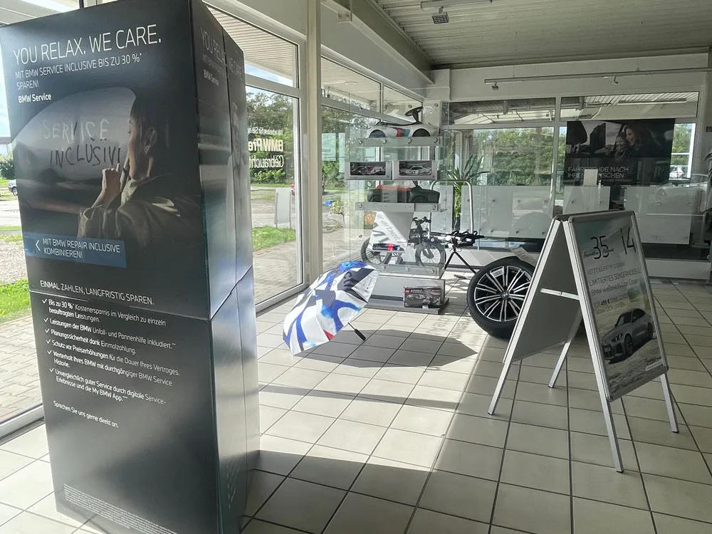 BMW-Servicewerbung in einem hellen Ausstellungsraum mit großen Fenstern, Regenbogen-Regenschirm, Fahrrädern und Werbetafeln.