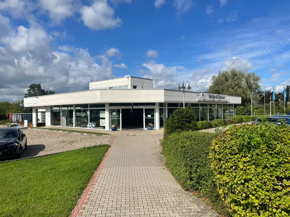 Gebäude mit großen Glasfenstern und einem Schild mit der Aufschrift Wolter & Steiner, umgeben von grünem Rasen und Sträuchern unter einem bewölkten blauen Himmel.