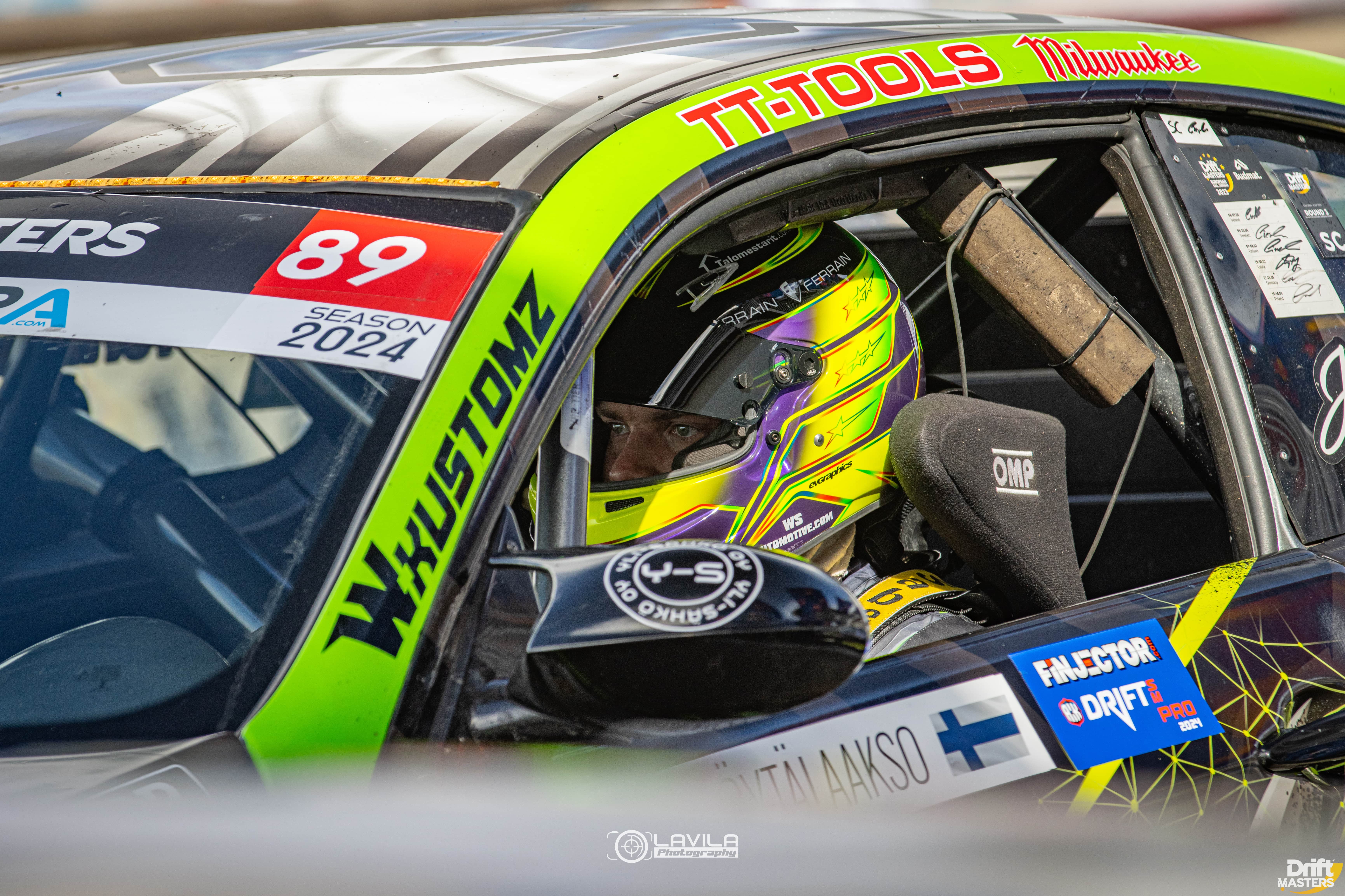 Race car driver wearing a yellow and purple helmet sitting inside a green and black drift car with racing stickers and safety harness.