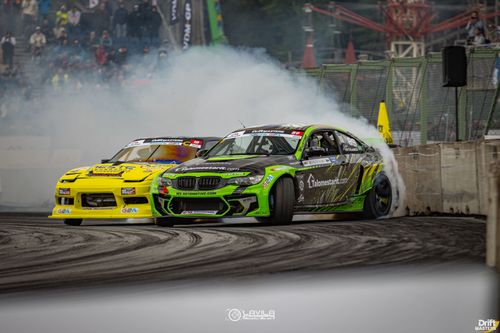 Two race cars drifting closely on a track, one green and black and the other bright yellow, with smoke trailing behind.