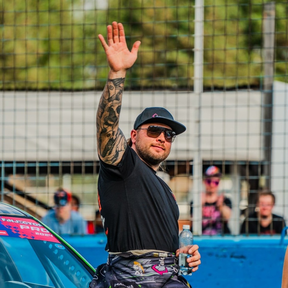 Man wearing sunglasses and a cap waves with a tattooed arm while holding a water bottle near a race car at a racing event.