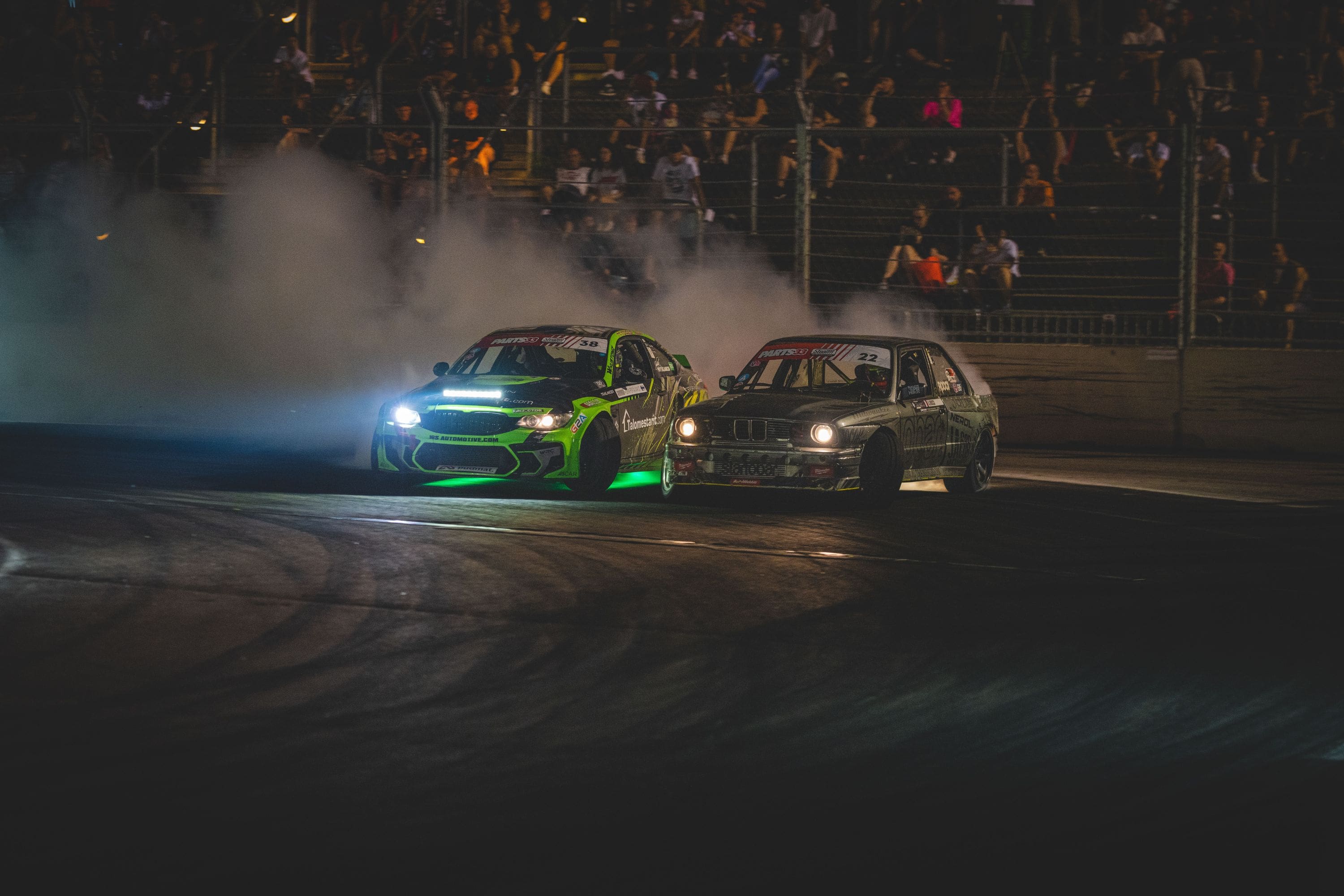 Two race cars drifting closely on a track at night with smoke and spectators in the background.