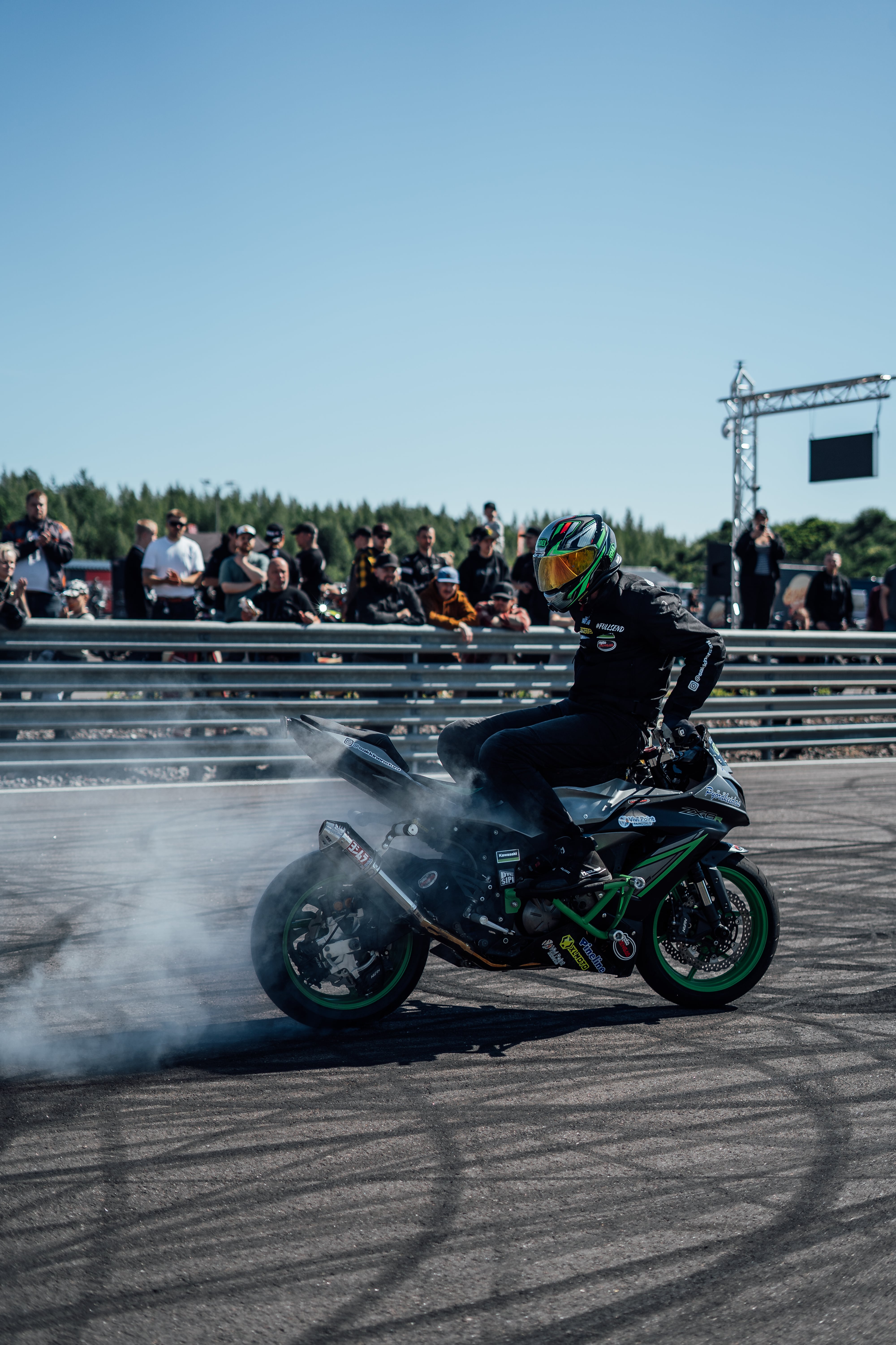 Motorcycle rider performing a burnout with smoke coming from the rear tire on a track, spectators watching behind a barrier.